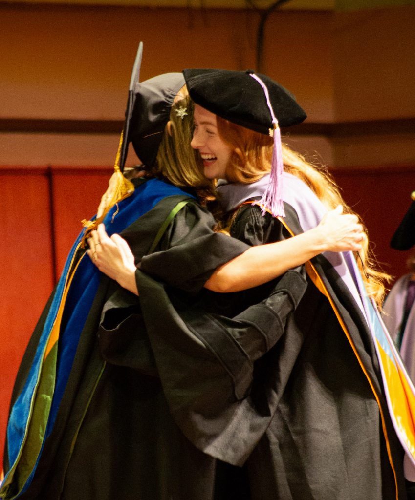 A School of Dentistry faculty member dressed in academic regalia hugs a graduating School of Dentistry Doctor of Dental Surgery student dressed in doctoral regalia during the 2024 commencement ceremony. 