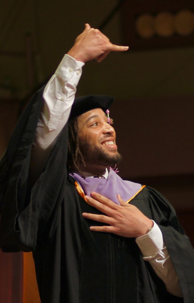 A graduating School of Dentistry Doctor of Dental Surgery student wearing doctoral regalia signs 'I love you' in American sign language to the crowd while walking across the stage at the 2024 commencement ceremony.