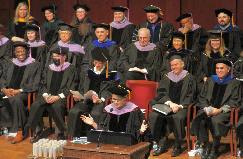 Laurie McCauley wears academic regalia while standing at a podium on a stage and speaking to an audience during the 2024 commencement ceremony. 