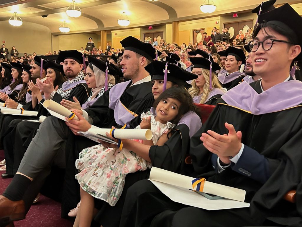 A graduating School of Dentistry Doctor of Dental Surgery student sits among fellow graduating Doctor of Dental Surgery students dressed in doctoral regalia in the audience with her daughter after receiving a diploma at the School of Dentistry 2024 commencement ceremony. 