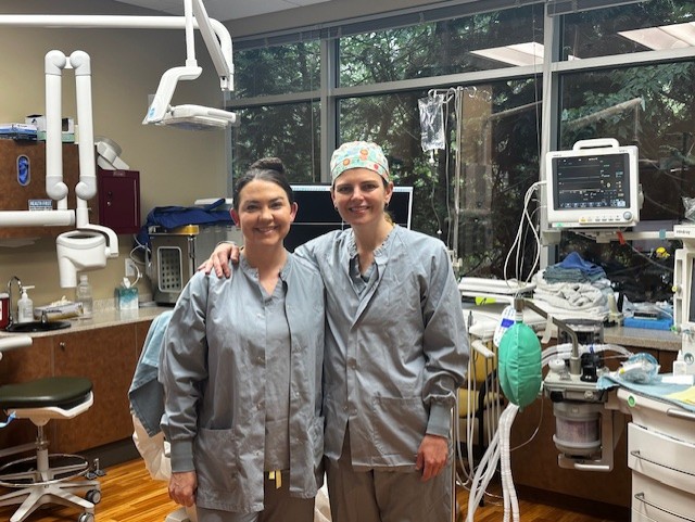 Two dentists pose in a dental treatment room while wearing scrubs. 